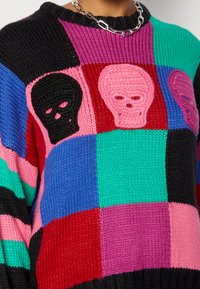 Maglione lavorato a maglia con un motivo a patchwork colorato che presenta sezioni nere, rosa, rosse, verdi e blu, e motivi di teschi stilizzati in nero e rosa.