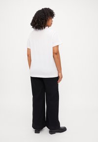 Weißes Baumwoll-T-Shirt mit lässiger Passform, kurzen Ärmeln und rundem Ausschnitt, kombiniert mit hochgeschnittenen schwarzen weit geschnittenen Hosen und Stiefeletten mit Blockabsatz.