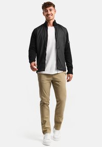 Sort bomberjakke med ribbede manchetter, lynlåse og kontrastfarvede ærmer, parret med en hvid T-shirt og beige bukser. Hvide sneakers fuldender looket.