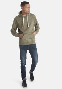 Felpa verde oliva con cappuccio e coulisse, tasche a marsupio, polsini a coste, abbinata a jeans slim-fit blu scuro e sneakers nere.
