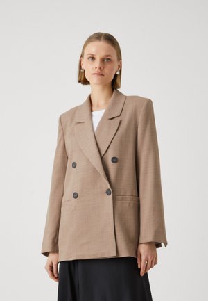Beige doppelt-knöpfbarer Blazer aus strukturiertem Stoff, mit schwarzen Knöpfen, Reverskragen und Seitentaschen, über einem weißen Oberteil getragen.