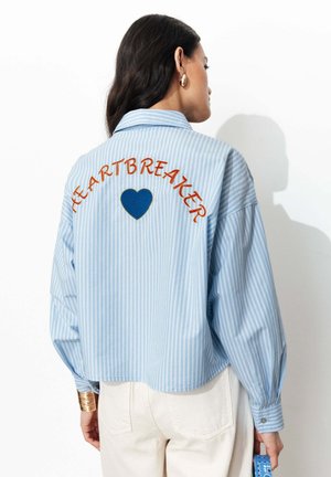 Femme portant une chemise bleu clair à fines rayures avec le texte « HEARTBREAKER » et un écusson cœur bleu dans le dos, associée à un pantalon blanc.