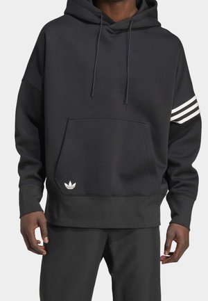 Homme portant un sweat à capuche noir avec une poche devant et trois bandes blanches sur la manche gauche, assorti à un pantalon noir.