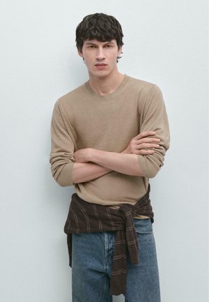 Giovane uomo con capelli scuri che indossa un maglione beige, jeans blu e una camicia a quadri scura legata intorno alla vita, in piedi con le braccia incrociate.