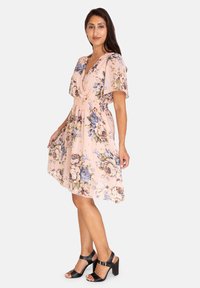 Blumenkleid aus hellpinkem Stoff mit blauen und gelben Blumen, tiefem V-Ausschnitt, elastischem Bund und kurzen, schwingenden Ärmeln. Schwarze Sandalen mit Blockabsatz.