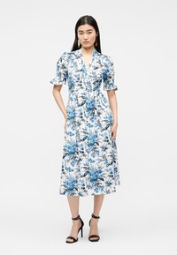 Vrouw draagt een witte midi-jurk met blauw bloemenprint, korte pofmouwen en zwarte enkellaarsjes met hoge hak en enkelbandje, staand tegen een effen achtergrond.