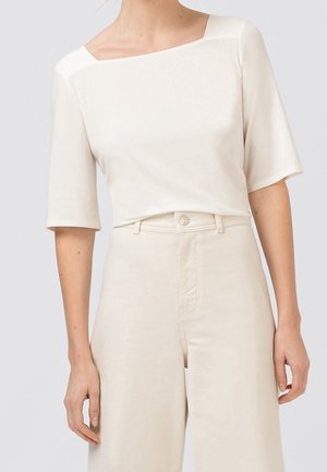 Mujer que lleva un top color crema con cuello cuadrado y mangas hasta el codo, combinado con pantalones anchos de tiro alto en color crema.