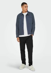 Camicia blu a maniche lunghe con due tasche sul petto, t-shirt bianca sotto, pantaloni neri e sneakers bianche. Fit slim, design casual.