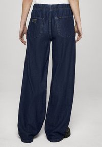 Platus platėjantys džinsai tamsiai mėlynos spalvos, su elastinga juosmens juosta ir dviem užpakalinėmis kišenėmis su etiketės akcentais. Lygi tekstūra be raštų.