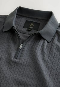 Polo gris ajustado con un patrón de diamante texturizado, cierre de cremallera en el cuello y cuello acanalado. La etiqueta dice "NEXT" y talla Mediana.