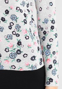 Grå kortkorts tröja med ett blommönster av svarta, blå och rosa blommor, åtföljd av gröna blad på ett mjukt, strukturerat tyg.