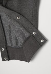 Dunkelgraues Kleidungsstück mit einem weichen, flauschigen Innenfutter, Metalldruckknöpfen und einem gerippten Bunddetail. Das Material wirkt strukturiert und warm.