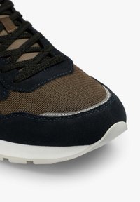 Primer plano de la punta de una zapatilla marrón y negra con cordones negros, tela de malla, ribete de ante y suela de goma blanca sobre fondo blanco.