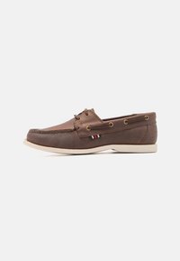 Zapato de barco de cuero marrón con suela de goma blanca, punta redondeada y ojales dorados. Presenta un pequeño acento a rayas cerca del talón.