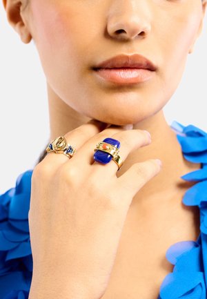 Bagues en or avec accents bleus et corail. L'une a une pierre bleue rectangulaire, l'autre présente un design de feuille texturé et plusieurs petites pierres précieuses.