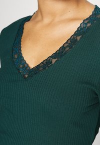 Tummanvihreä ribbipaita, jossa on V-mallinen kaula-aukko, joka on koristeltu monimutkaisella mustalla pitsireunuksella ja kukkakuviolla, korostaen tekstuuria ja yksityiskohtia.