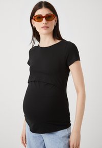 Črna materinska majica s kratkimi rokavi, ki ima plasten dizajn nad trebuhom. Gladko bombažno blago, okrogla izreza in sproščen kroj.