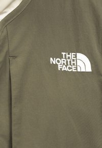 Ύφασμα σε ελιά πράσινο με λευκό κεντημένο λογότυπο "THE NORTH FACE" και σύμβολο μισού θόλου στην άνω περιοχή του στήθους ενός ενδύματος.