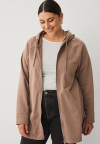 Brown zip-up hoodie with a hood, featuring a relaxed fit and textured fabric. Paired with a light top and dark jeans for contrast.