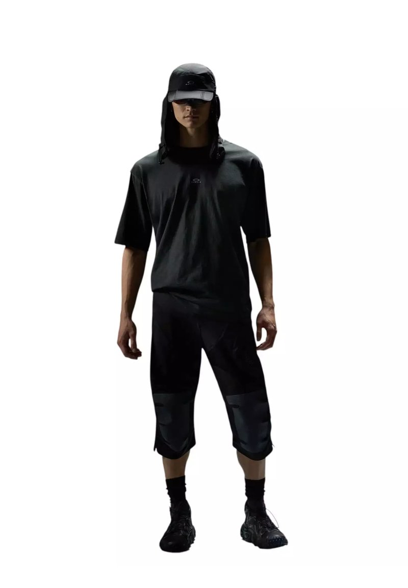 Modèle masculin portant une casquette noire avec rabats de cou, un t-shirt noir ample, un pantalon court noir avec des renforts aux genoux, des chaussettes noires et des baskets noires.