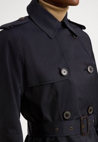 Καμπαρντίνα σε ναυτικό μπλε χρώμα με διπλή σειρά κουμπιών, επωμίδες και ζώνη. Κατασκευασμένη από λείο ύφασμα με ελαφρώς γυαλιστερή υφή, διακοσμημένη με χρυσές λεπτομέρειες.