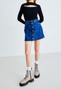 Svart långärmad topp med urklipp på ryggen, kombinerad med en blå jeanskjol med knappar framtill och cirkulära mönster, samt beige snörkängor.