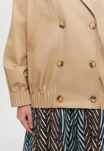 Beige dubbelzijdige jas met grote schildpadknopen, elastische manchetten en zijzak, gecombineerd met een rok met een multikleurig patroon.