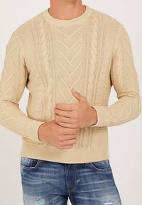 Beige kabelstickad tröja med ribbade muddar och fåll, bärs tillsammans med urtvättade blå slitna jeans, händerna knäppta vid midjan.