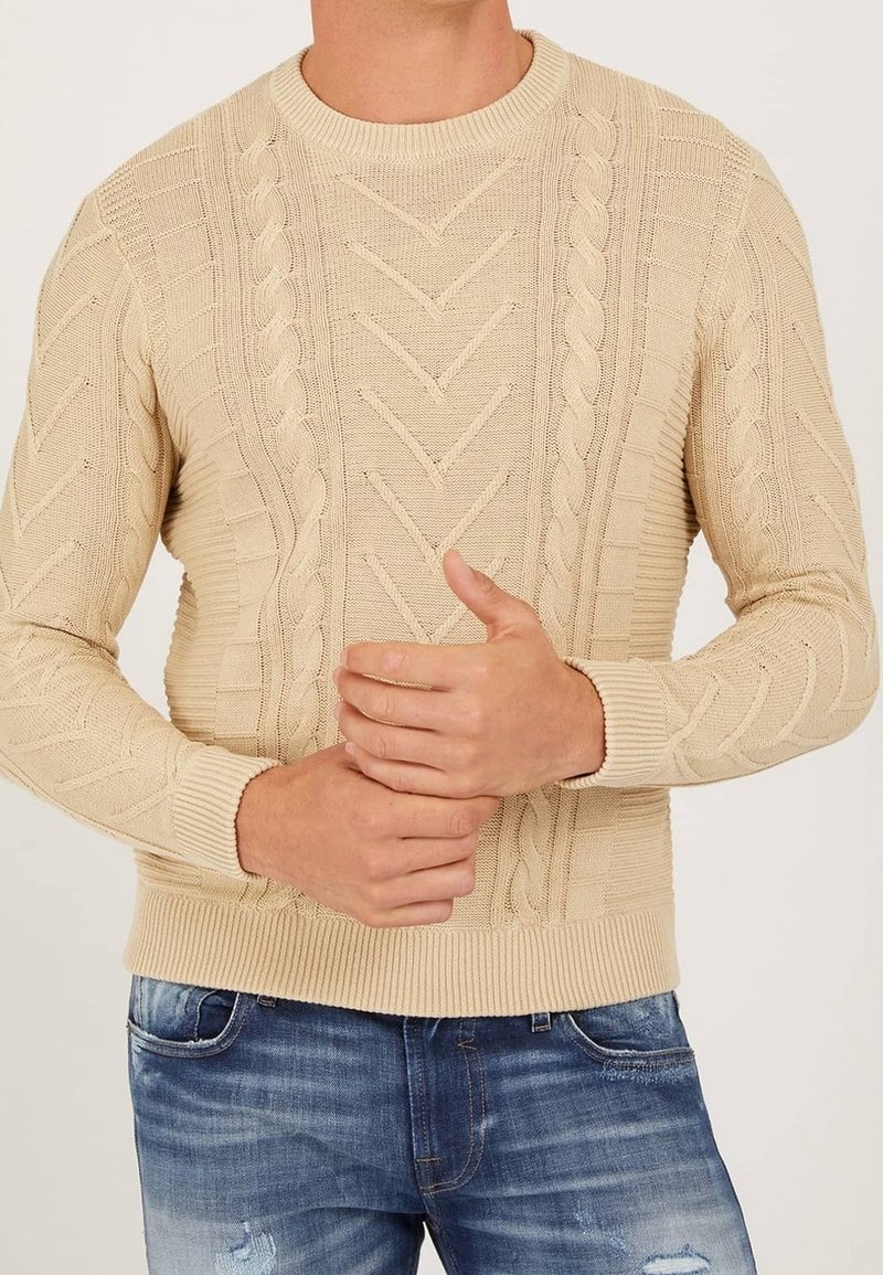 Beige kabelstickad tröja med ribbade muddar och fåll, bärs tillsammans med urtvättade blå slitna jeans, händerna knäppta vid midjan.