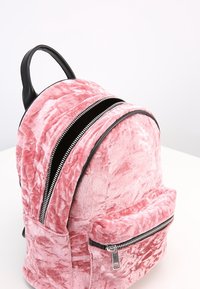 Mochila de terciopelo rosa con forma redondeada, detalles en cuero negro, dos compartimentos con cremallera y un bolsillo frontal con herrajes plateados.