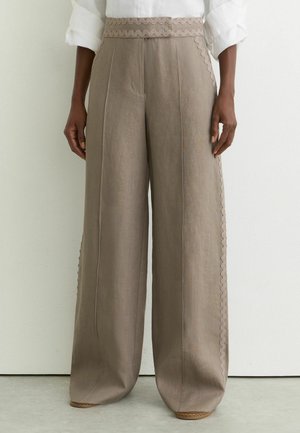 Beige wijde broek met zigzagzoom aan de taille en zijkanten, gedragen met een wit overhemd met opgerolde mouwen.