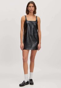 Schwarzes Leder-Minikleid mit quadratischem Ausschnitt, dünnen Trägern und glatter Textur. Kombiniert mit schwarzen Mokassins und weißen Socken.