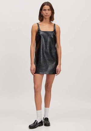 Zwarte leren mini-jurk met een vierkante halslijn, dunne bandjes en een gladde textuur. Gestyld met zwarte loafers en witte sokken.