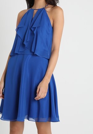 Bezrękawowa suknia w królewskim odcieniu niebieskiego z warstwową falbaną na gorsecie i zwiewną spódnicą w kształcie litery A, na osobie stojącej.