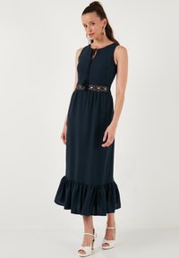 Navy blue pitkä mekko, jossa on tiukka vartalo, avonainen kaula-aukko, pitsiyksityiskohtia vyötäröllä ja liehuva helma. Valmistettu kevyestä kankaasta.
