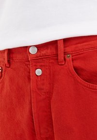 Røde denimbukser har høy midje, metallknapper og sømdetaljer. Stoffet har en lett teksturert utseende.