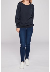 Sweat-shirt bleu marine foncé à col rond et poignets côtelés, associé à un jean bleu slim et des baskets blanches. Présente un petit logo sur la poitrine.