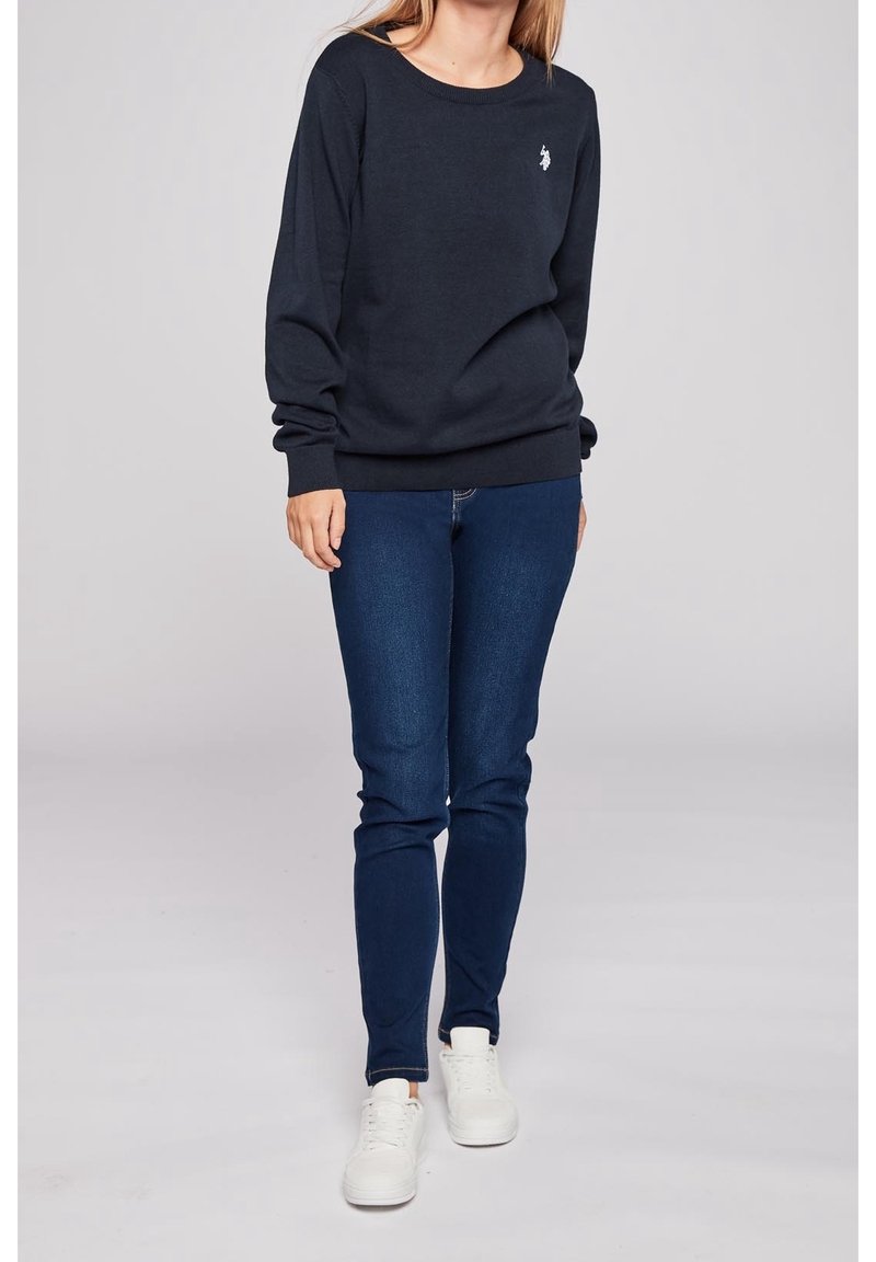 Sweat-shirt bleu marine foncé à col rond et poignets côtelés, associé à un jean bleu slim et des baskets blanches. Présente un petit logo sur la poitrine.