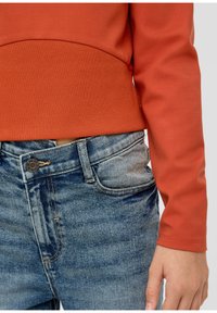 Roestkleurige sweatshirt met ribbelband, gedragen op lichtblauwe jeans met vervaging en een metalen knoopaccent. De stof lijkt zacht.