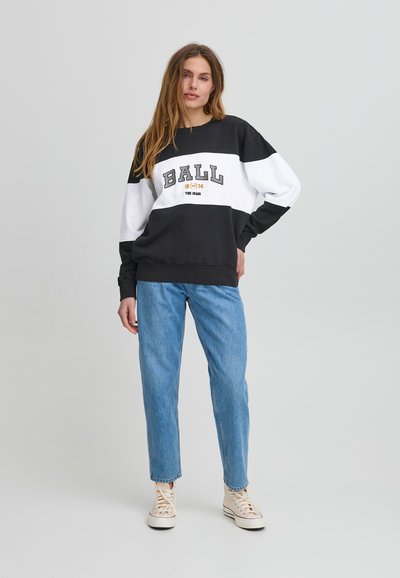 Sudadera en blanco y negro con "BALL 1974" en texto gris, combinada con jeans rectos de color azul claro y zapatillas beige.