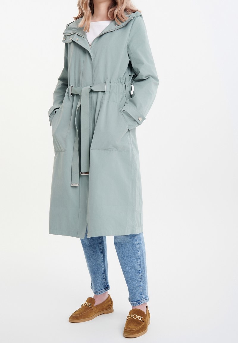 Světle zelený dlouhý trench coat s kapucí, výrazně stáhnutým pasem a dvěma předními kapsami. Nosič má na sobě džíny a hnědé semišové mokasíny.
