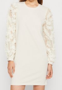 Roomkleurige jurk met lange doorschijnende mouwen, versierd met bloemapplicaties, en een ronde halslijn. Gladde stof met geribbelde manchetten aan de mouwen.