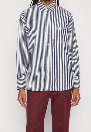 Zwart-wit verticaal gestreept overhemd met knopen en een borstzak, gecombineerd met bordeauxrode broek, gedragen door een persoon met een donkere huid en gevlochten haar.