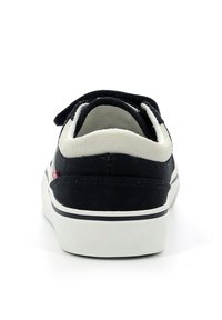 Zapatilla de lona negra con suela de goma blanca, que presenta un cuello acolchado y un acento de logo rojo. Cinta de velcro para un ajuste seguro.