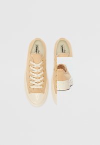 Beiga canvas sneakers med en glänsande krämfärgad tåhätta, gyllene öljetter och vita snören; bakvy visar en texturerad panel och en vit gummisula.
