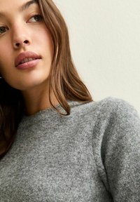 Pull en laine grise avec un col rond, présentant une texture douce et des motifs subtils, partiellement visible sur un modèle devant un fond neutre.