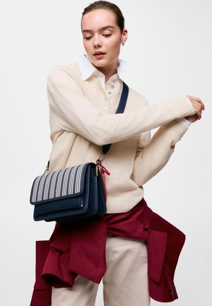 Femme portant un pull crème et une chemise blanche, tenant un sac bandoulière rayé bleu marine avec une veste bordeaux nouée autour de la taille.