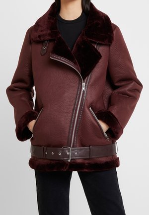 Kvinde iført en mørk burgundy fåreskindjakke med bred krave, diagonalt frontlynlås, lynlåslommer, pelsærmer og bælte i taljen.
