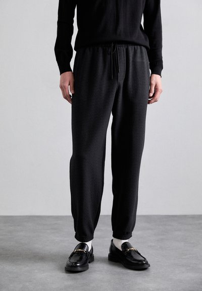 Schwarze, strukturierte Jogginghose mit einem elastischen Bund und Kordelzug. Kombiniert mit schwarzen Slippern und einem schwarzen Langarmshirt.