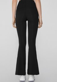 Leggings noirs évasés en matériau extensible avec taille haute. Présente une texture lisse et une silhouette ajustée, associés à des chaussures blanches.